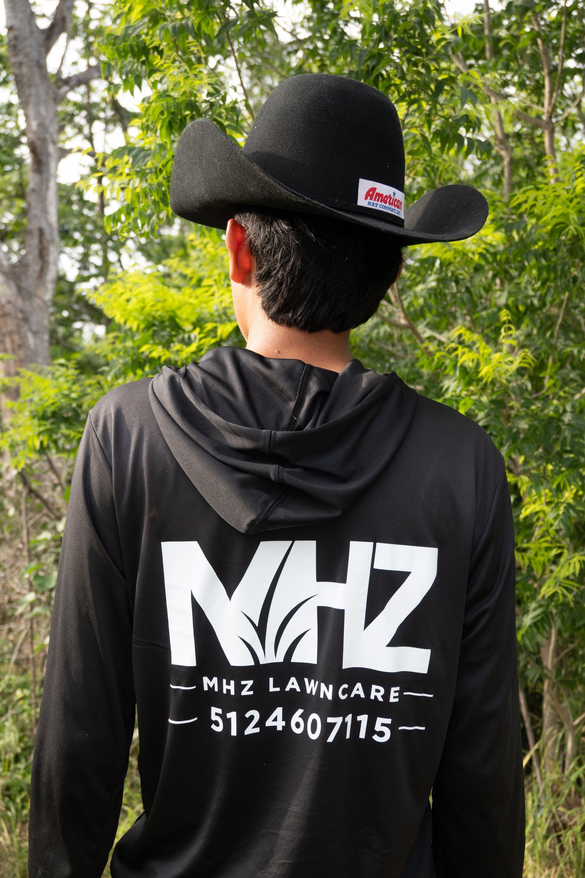 Person wearing black MHZ Lawncare hoodie and cowboy hat facing away, standing in front of trees