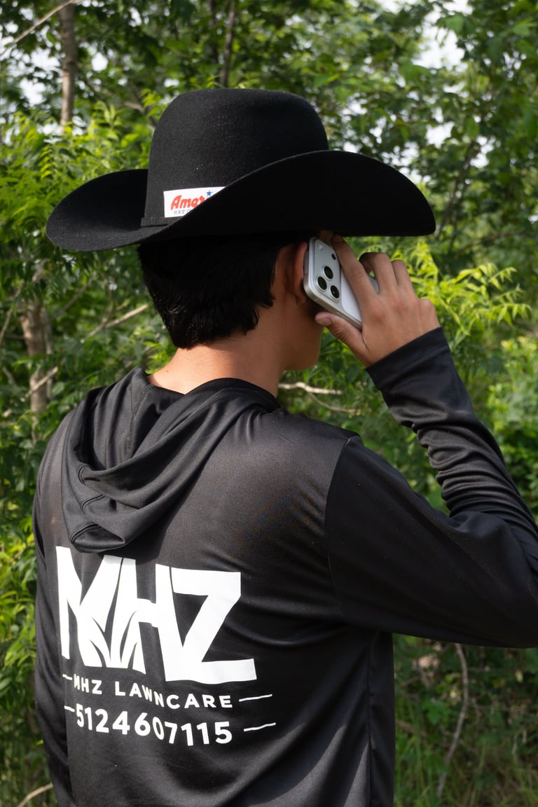 Person wearing black cowboy hat and branded hoodie taking a photo with a smartphone outdoors among trees