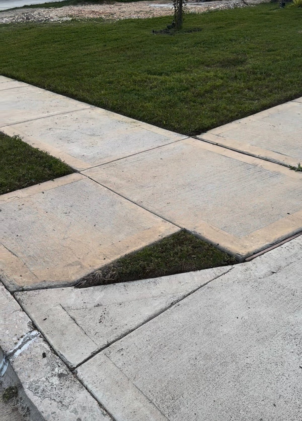 Uneven concrete sidewalk with a sunken section creating a tripping hazard, adjacent to green grass lawn