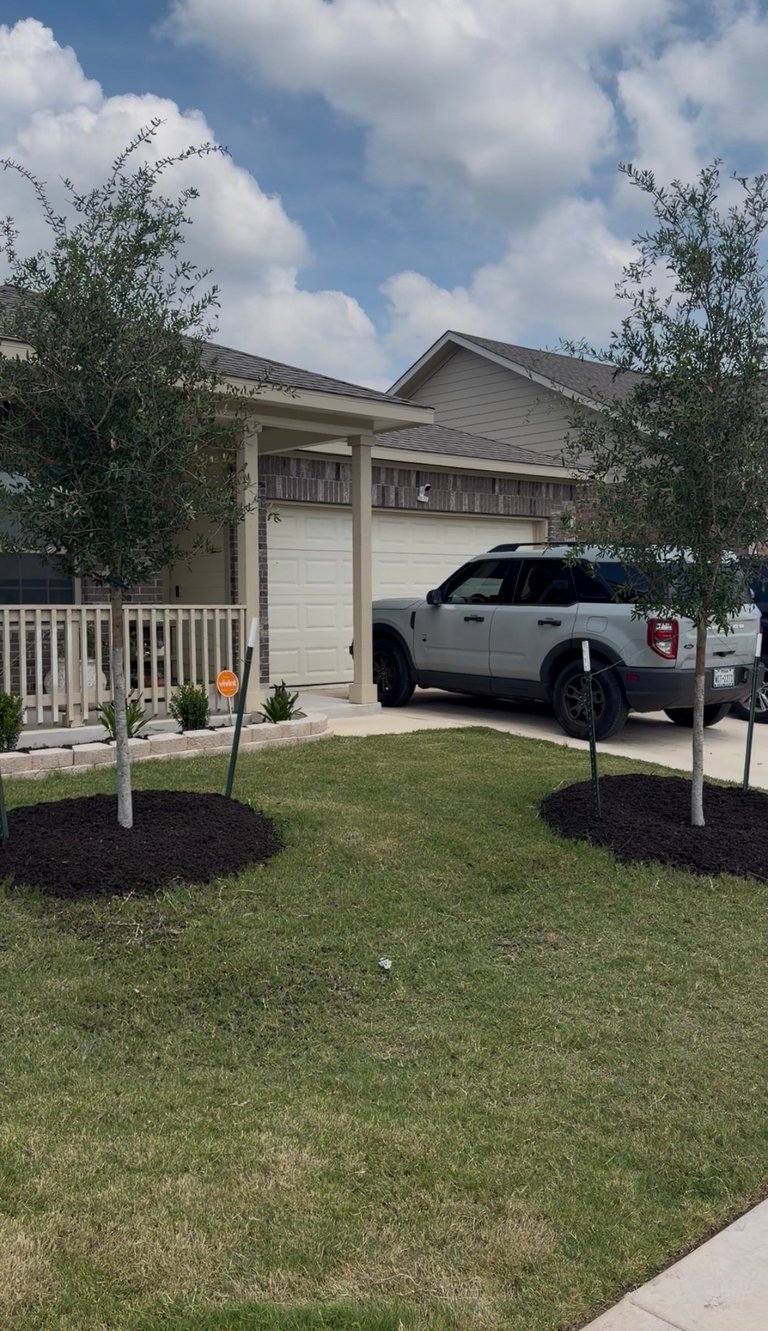Single-story home with white SUV in driveway, two-car garage, manicured lawn, and green trees under blue sky with clouds