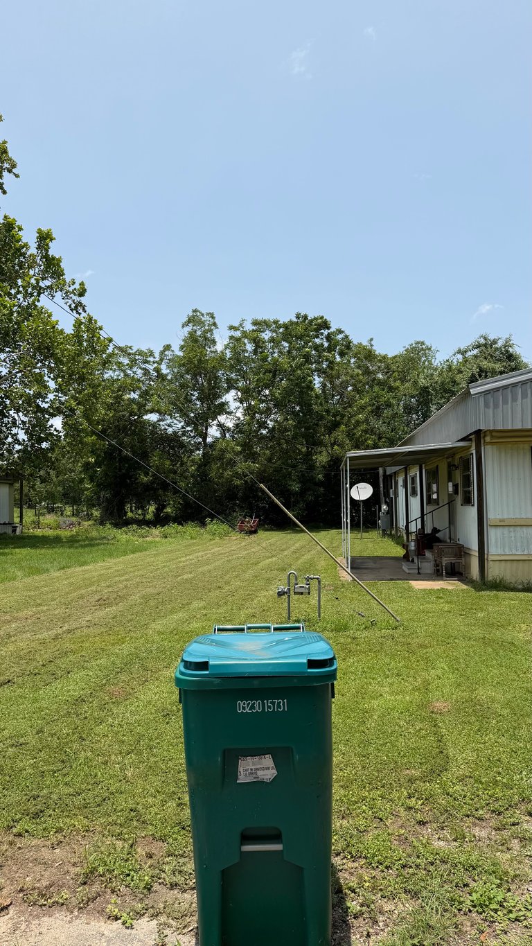 Green trash can with antenna in rural yard facing white metal house and trees under blue sky