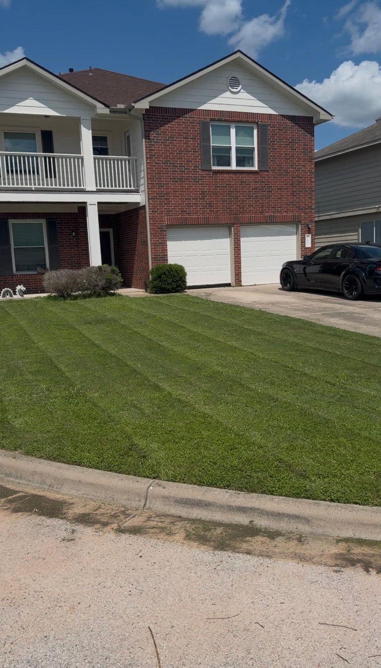 Two-story brick house with white garage doors, front porch, and manicured lawn with black car in driveway on a sunny day