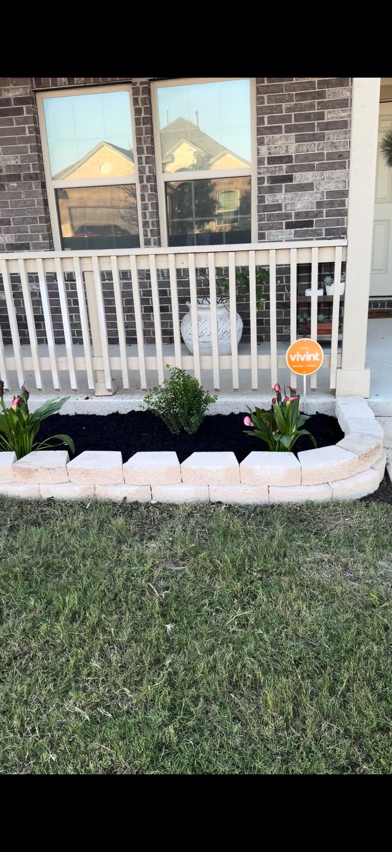 Front porch with white railing, brick exterior, and curved flower bed with black mulch and green plants